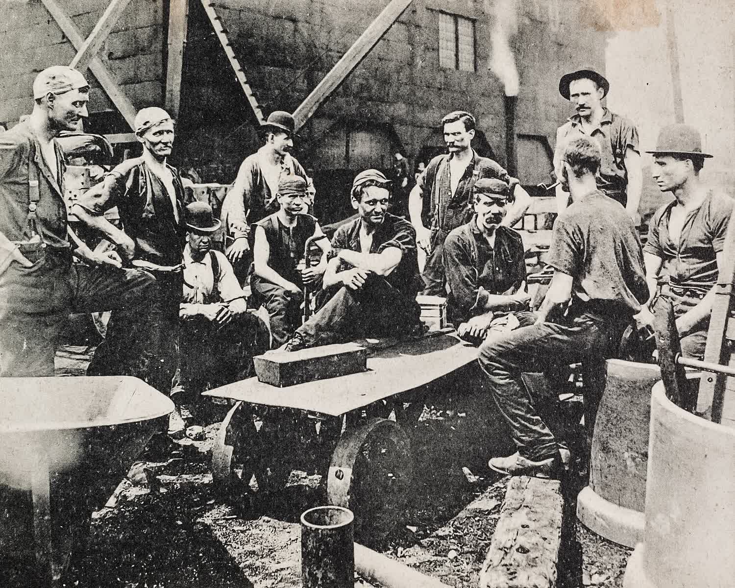 Fotografía de archivo de 1907: un grupo de trabajadores de una planta siderúrgica, sentados y de pie en su descanso de comida, con la nave industrial al fondo